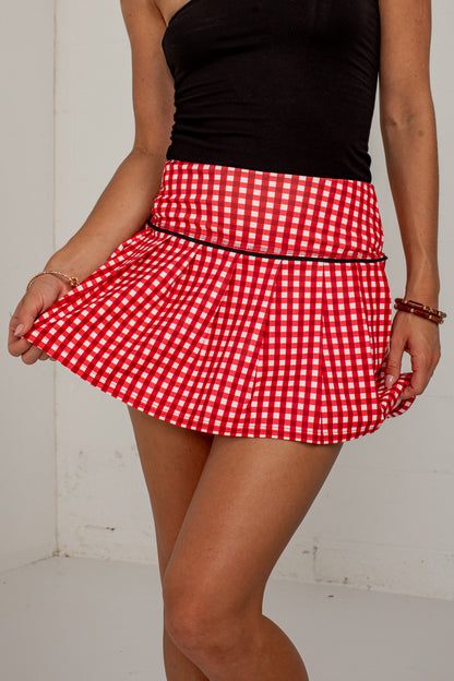 A red and white gingham pleated tennis skirt with a thin black waistband, modeled with a black strapless top against a white wall.
