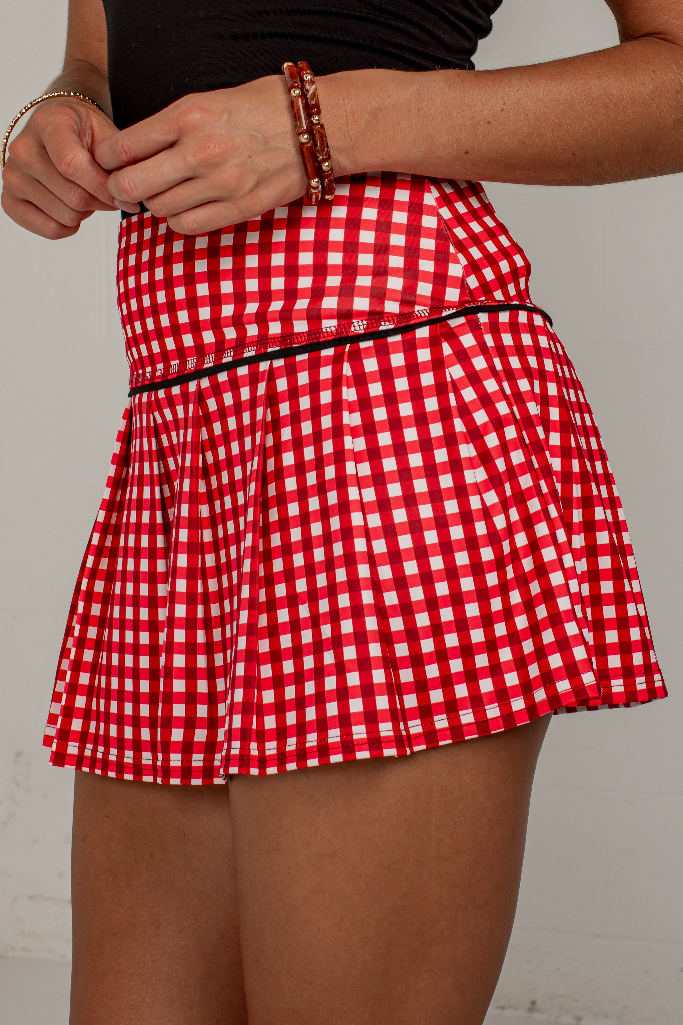A red and white gingham pleated tennis skirt with a thin black waistband, shown on a model against a light gray background.