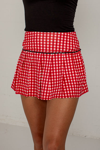 A red and white gingham pleated mini skirt with a thin black waistband trim, shown on a model wearing a black top against a light gray wall.