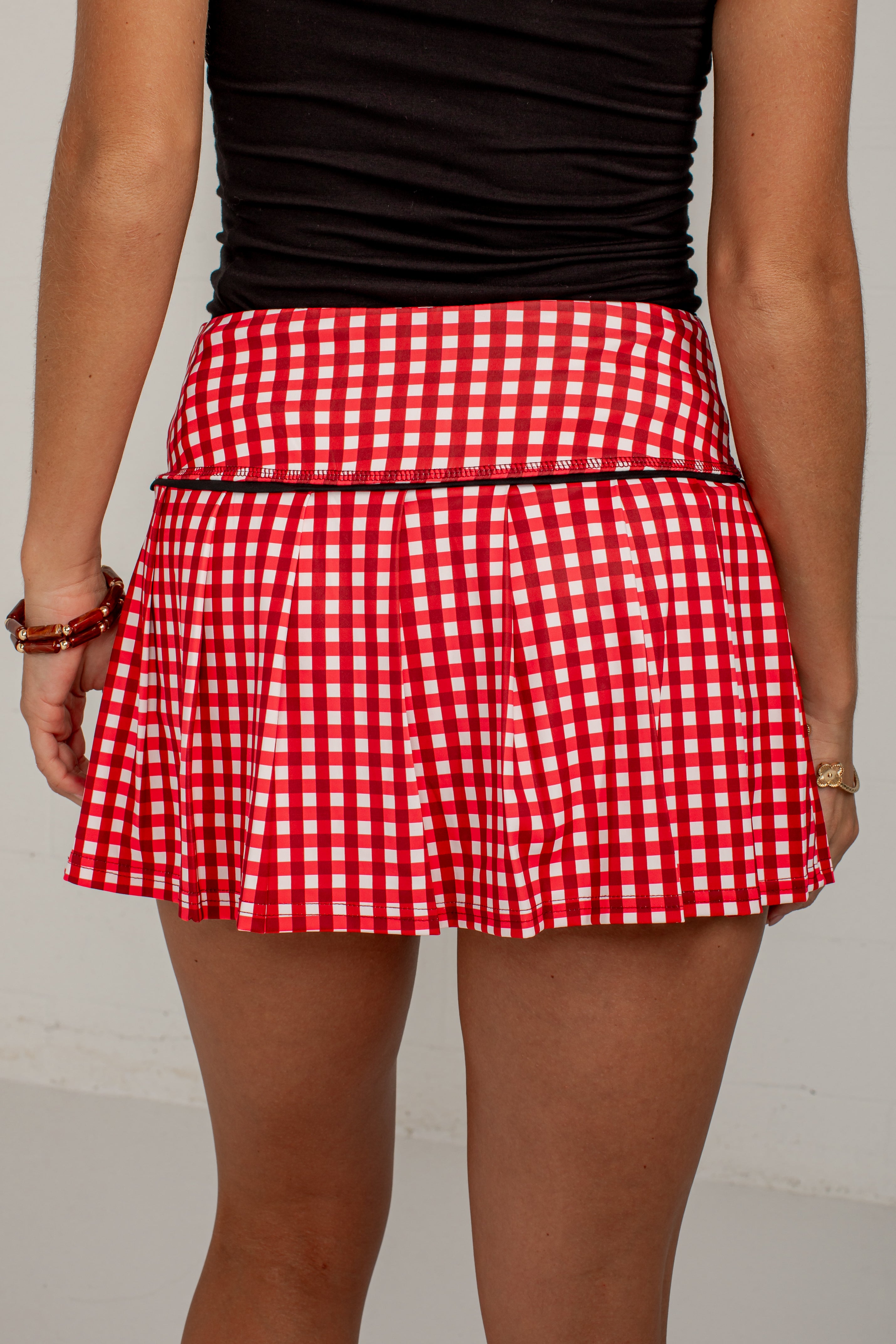 A red and white gingham pleated tennis skirt with a thin black waistband trim, shown on a model from the back.