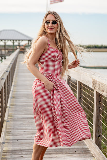 Woman in a red checkered dress standing on a wooden pier by a body of water.