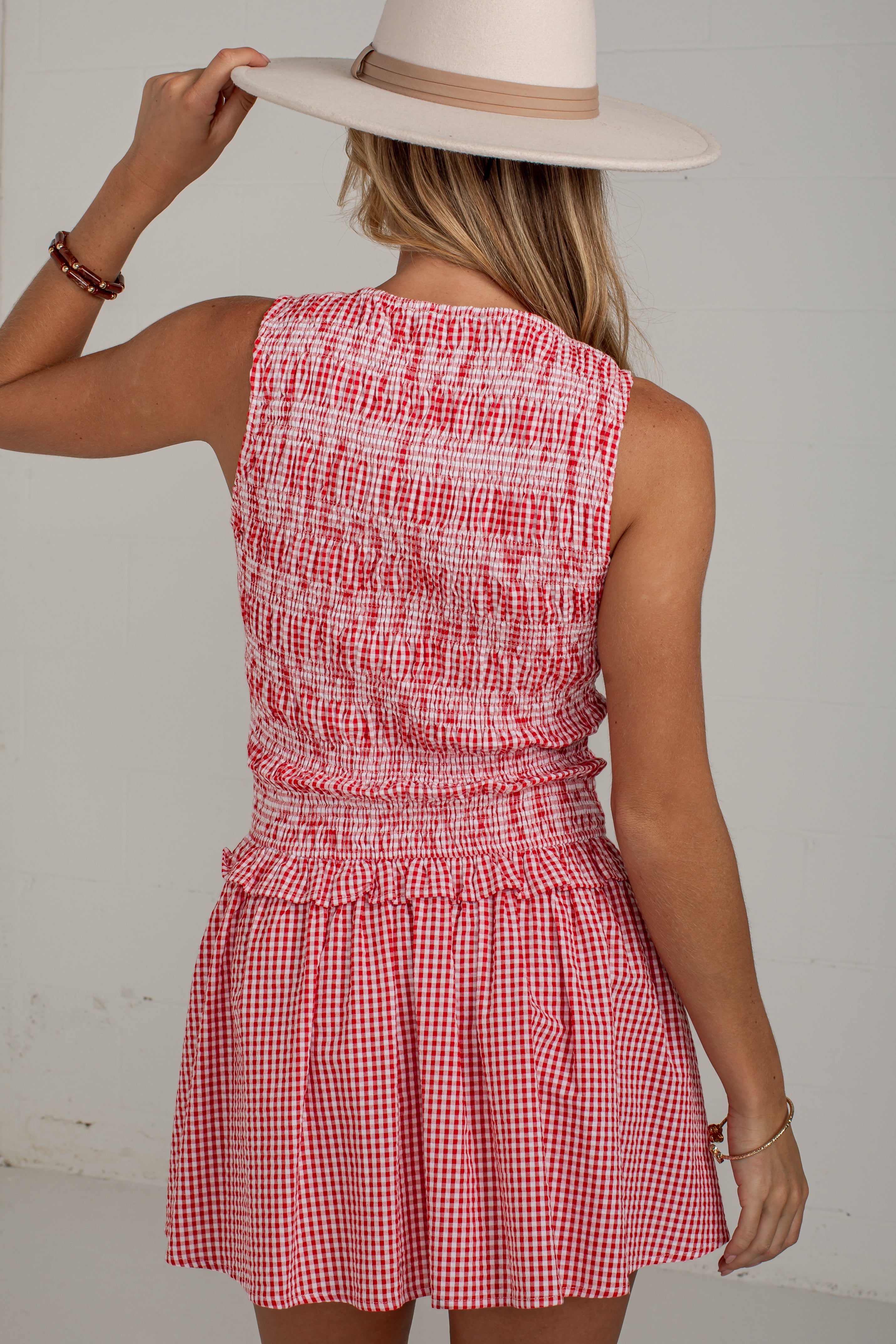 A woman shown from the back wearing a sleeveless red and white gingham mini dress with a smocked bodice and a wide-brimmed cream hat, set against a plain light gray wall.