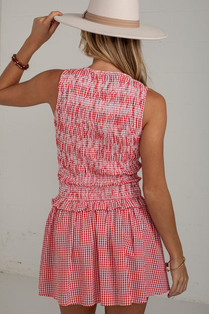 A woman shown from the back wearing a sleeveless red and white gingham mini dress with a smocked bodice and a wide-brimmed cream hat, set against a plain light gray wall.