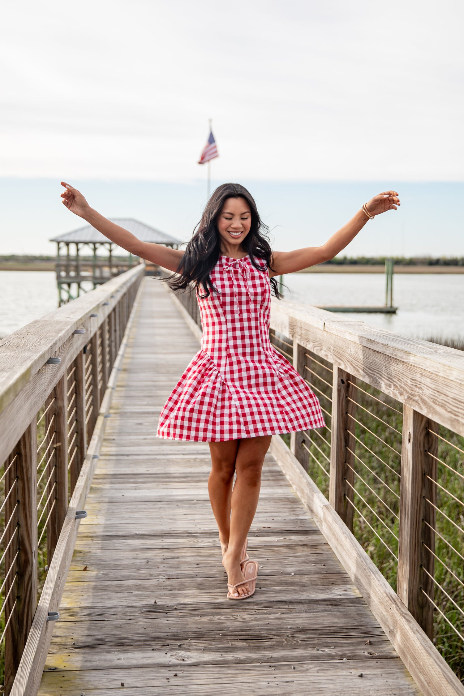 Woman in a red and white checkered dress standing on a wooden dock by a lake.