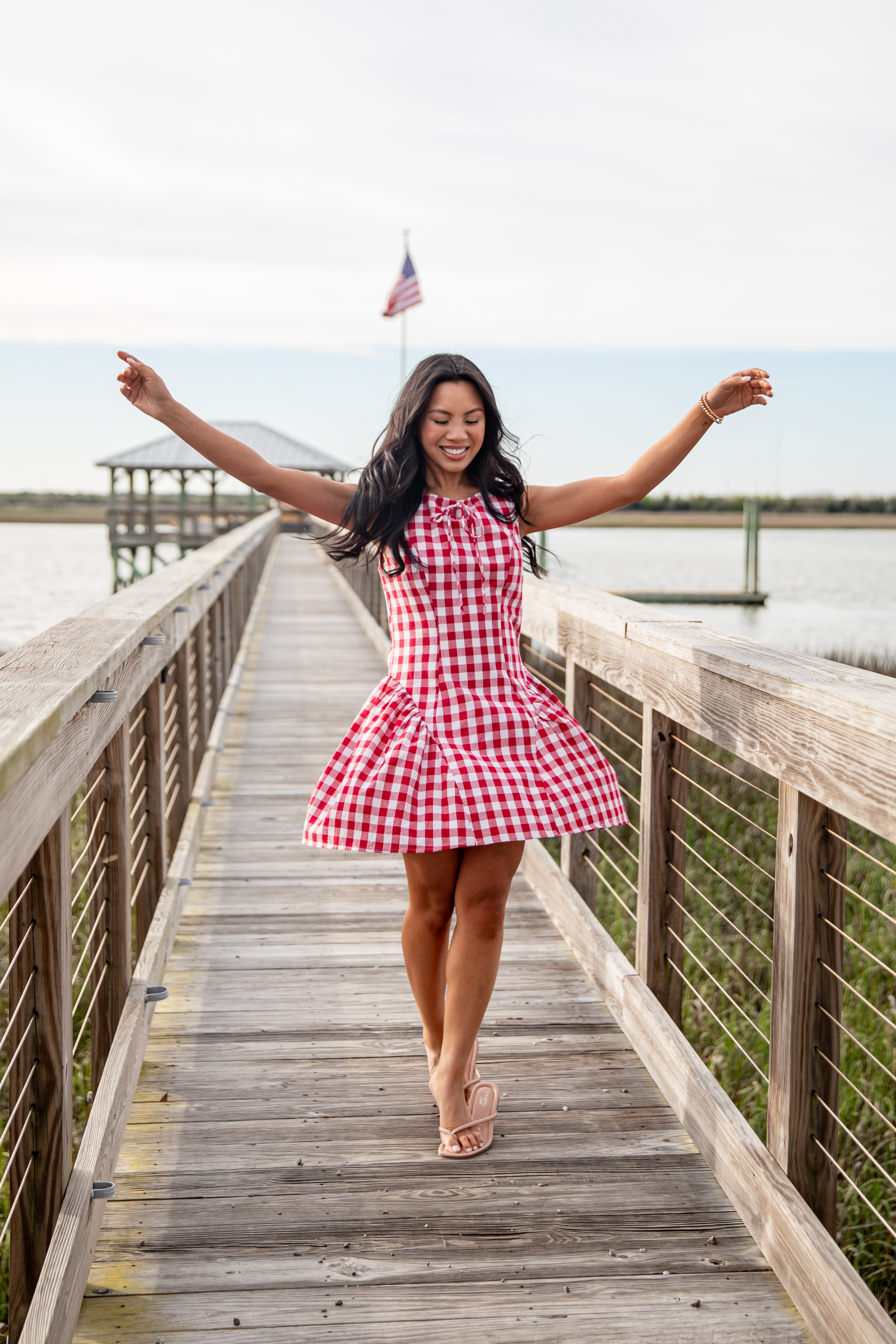 Woman in a red and white checkered dress standing on a wooden dock by a lake.