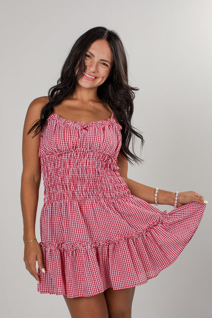 A woman wearing a red and white gingham mini dress with a smocked bodice and ruffled straps, posing against a light gray studio background.