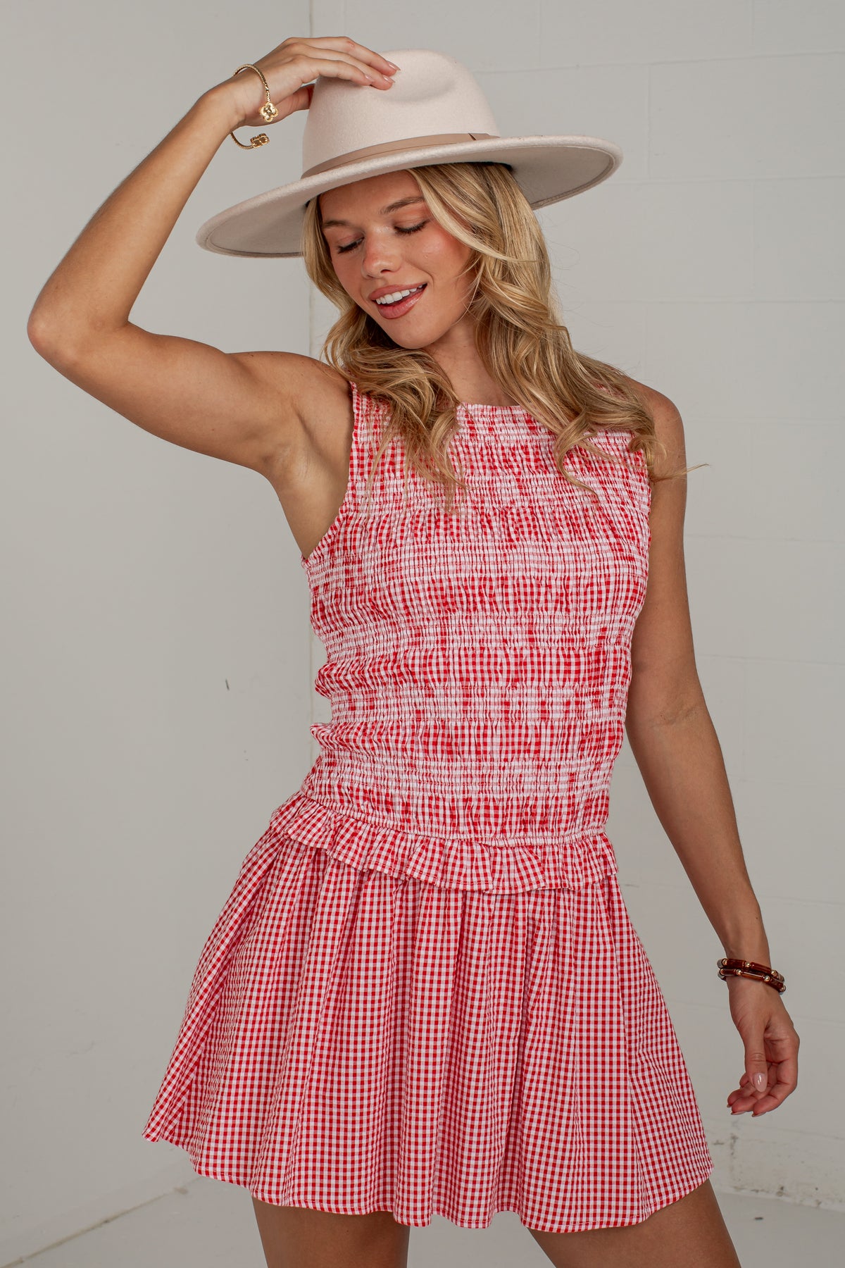 A woman wearing a red and white gingham sleeveless dress with a smocked bodice and a wide-brimmed light beige hat, posing against a plain light gray wall.
