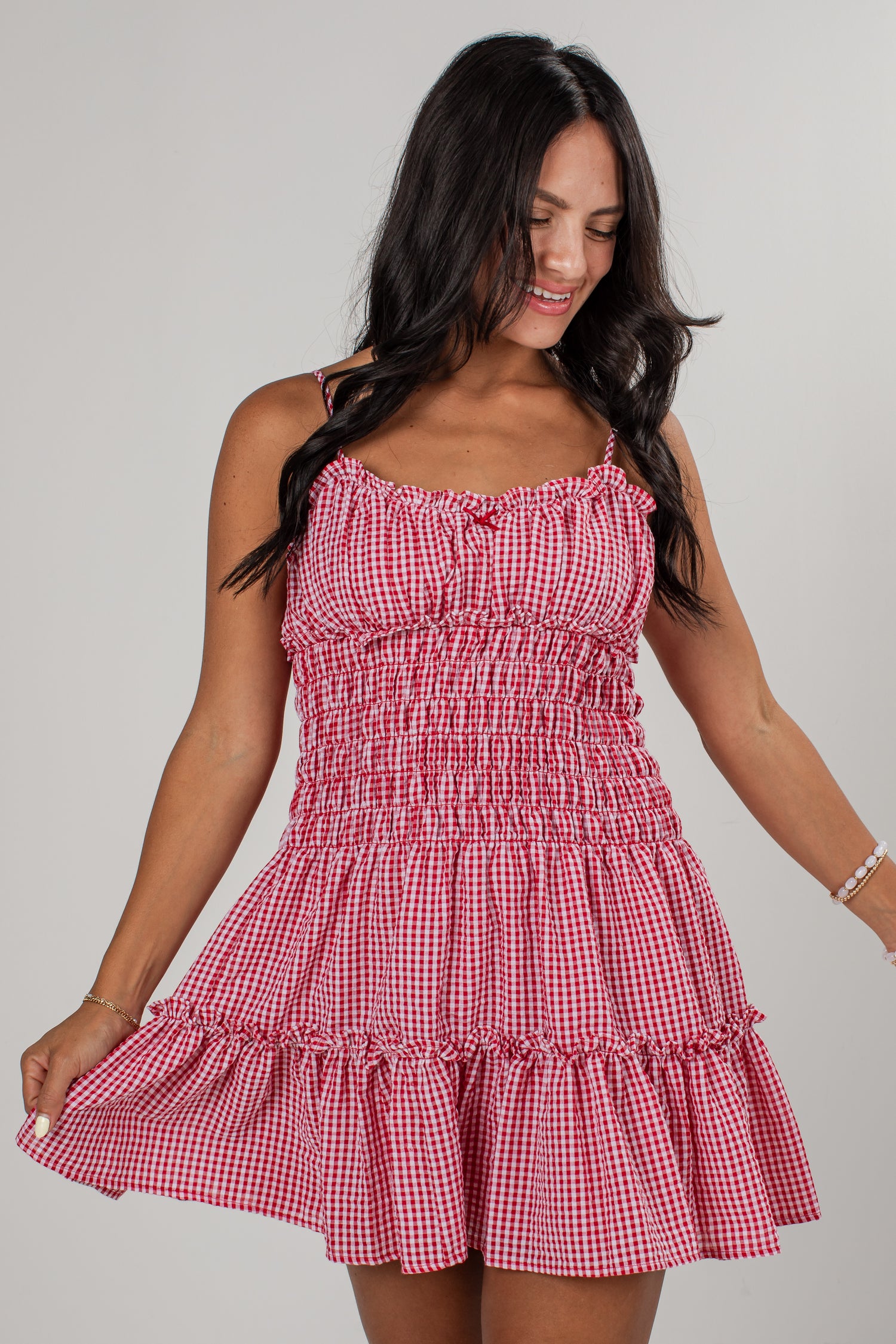 A woman wearing a red and white gingham mini dress with a smocked bodice and ruffled straps, posing against a light gray studio background.