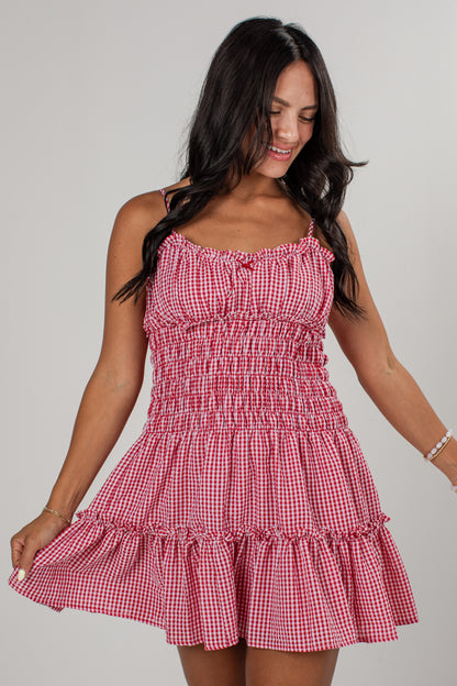 A woman wearing a red and white gingham mini dress with a smocked bodice and ruffled straps, posing against a light gray studio background.