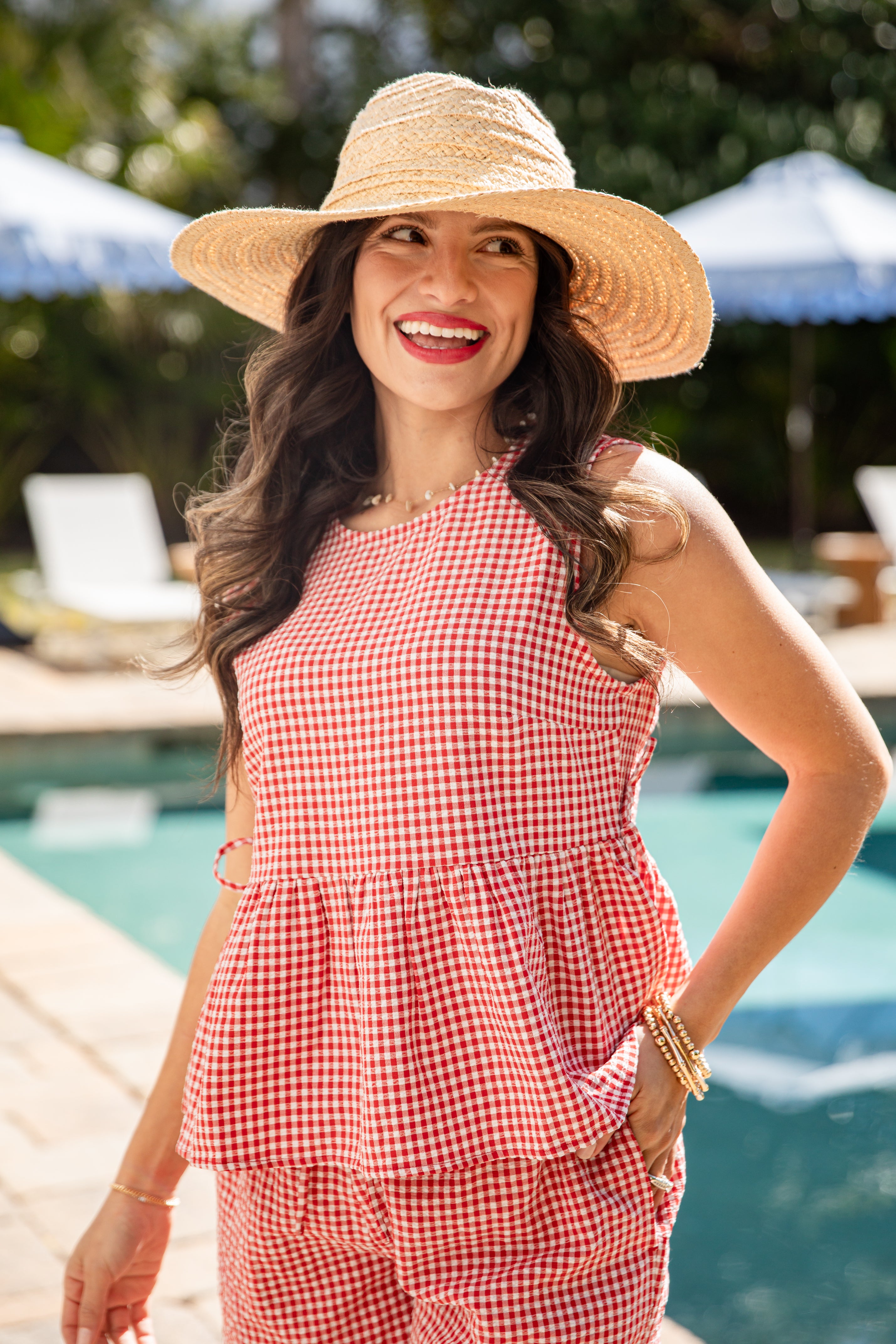 Woman wearing a red checkered outfit and straw hat by a poolside.