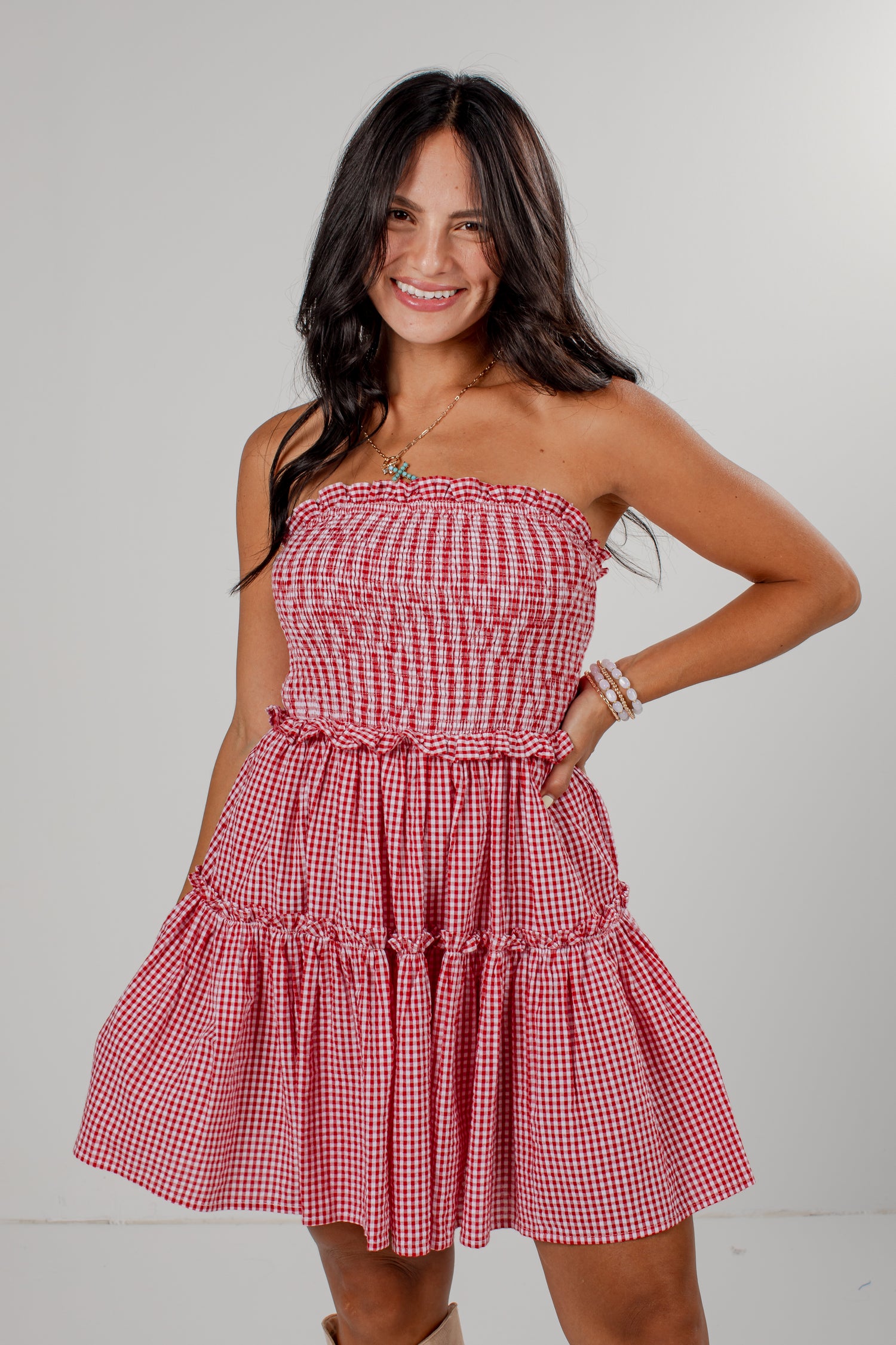 A woman modeling a red and white gingham strapless mini dress with a smocked bodice and tiered skirt, standing against a light gray studio background.