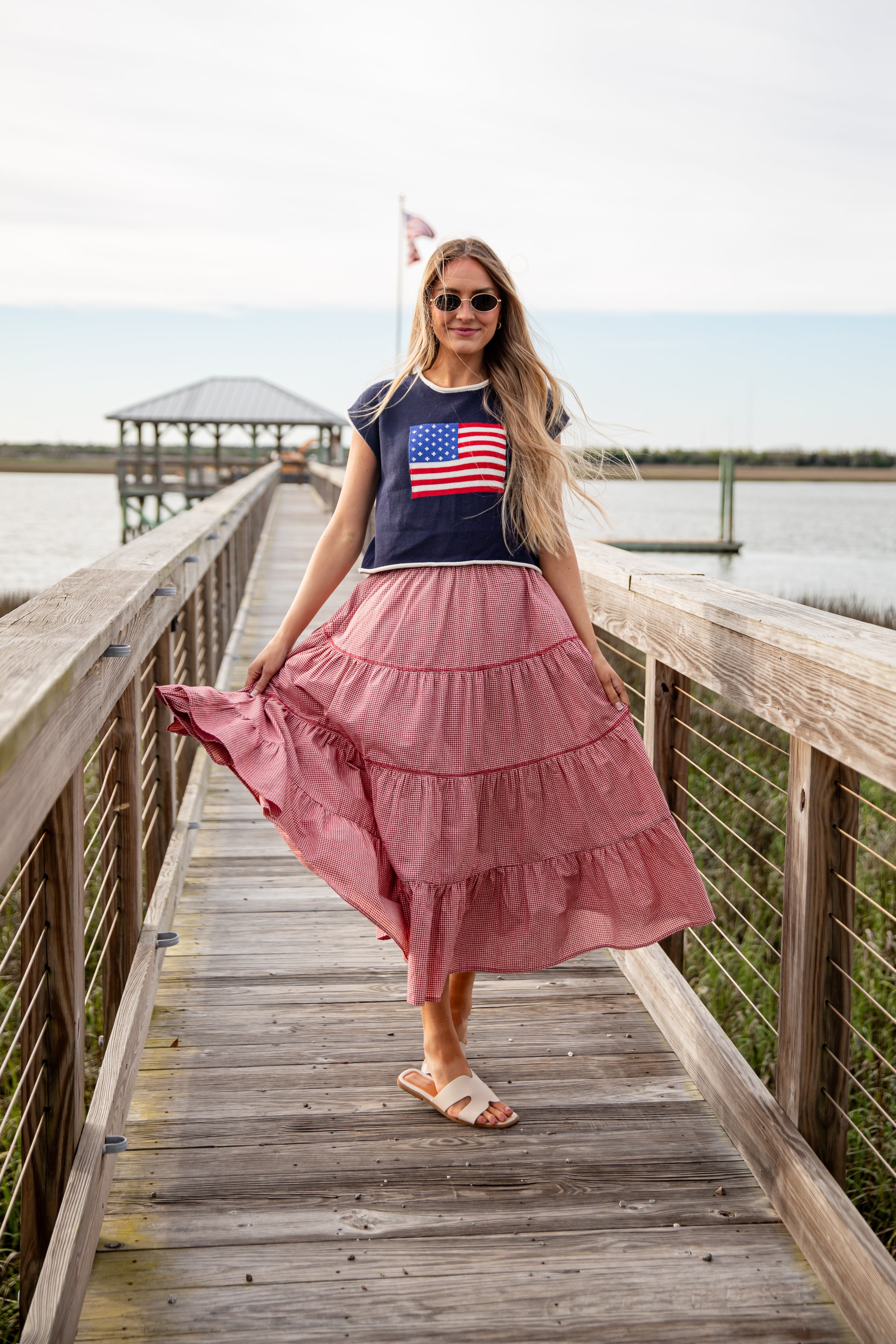 Woman in a pink skirt and American flag t-shirt standing on a wooden dock by a body of water.