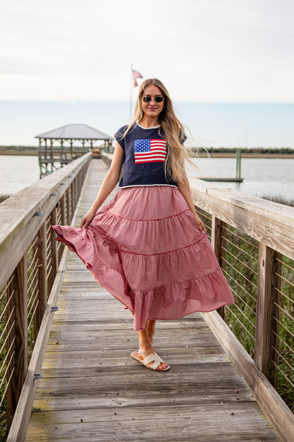 Woman in a pink skirt and American flag t-shirt standing on a wooden dock by a body of water.