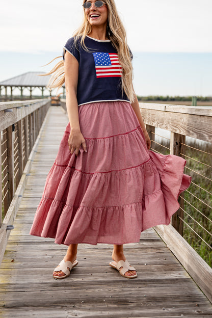 Woman wearing a navy t-shirt with an American flag design and a pink tiered skirt on a wooden boardwalk.