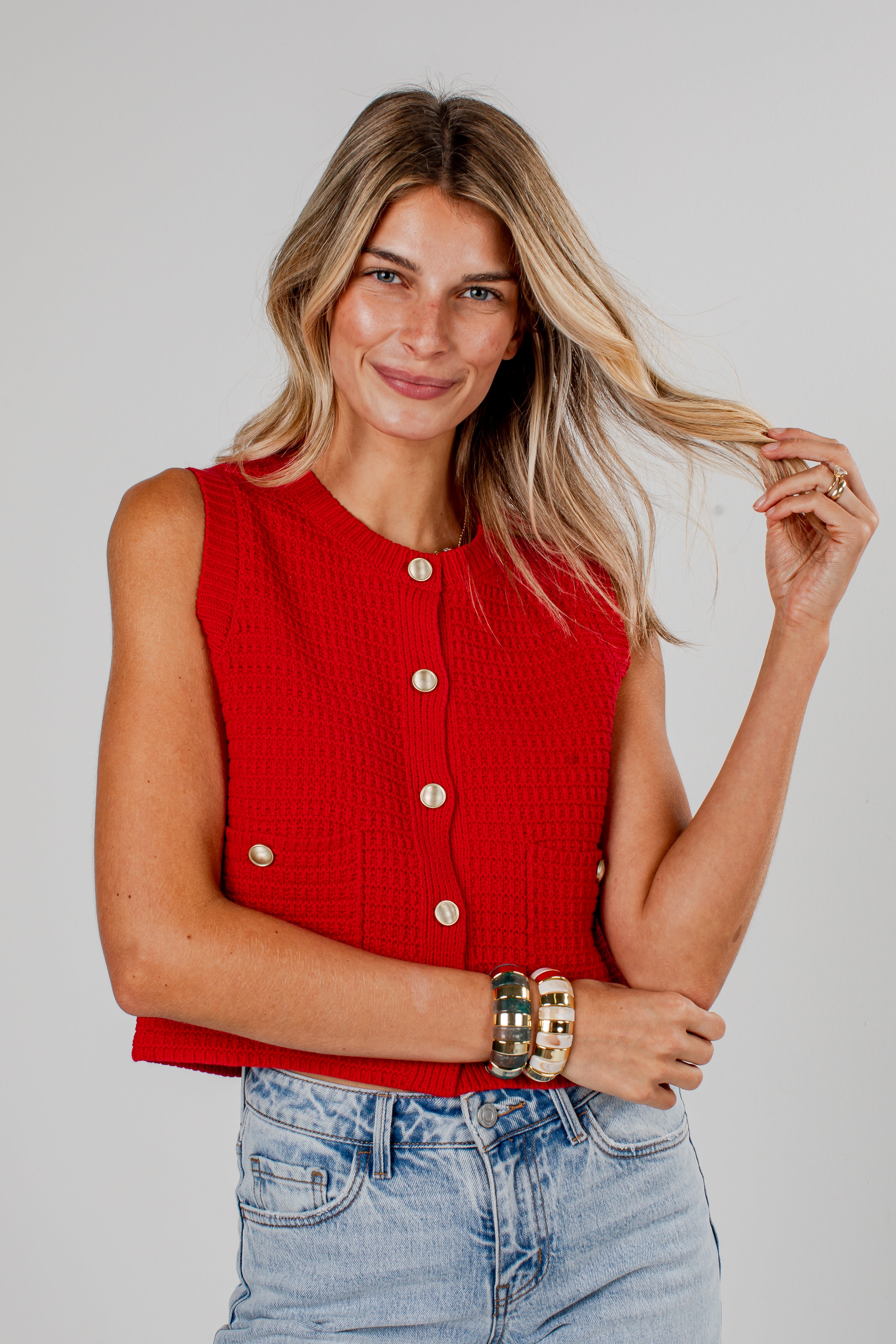 A woman modeling a sleeveless red knit vest with gold-tone buttons, paired with light-wash blue jeans, against a solid light gray background.