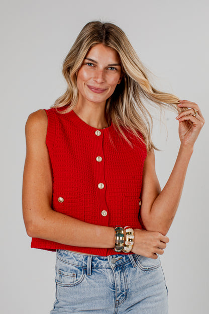 A woman modeling a sleeveless red knit vest with gold-tone buttons, paired with light-wash blue jeans, against a solid light gray background.