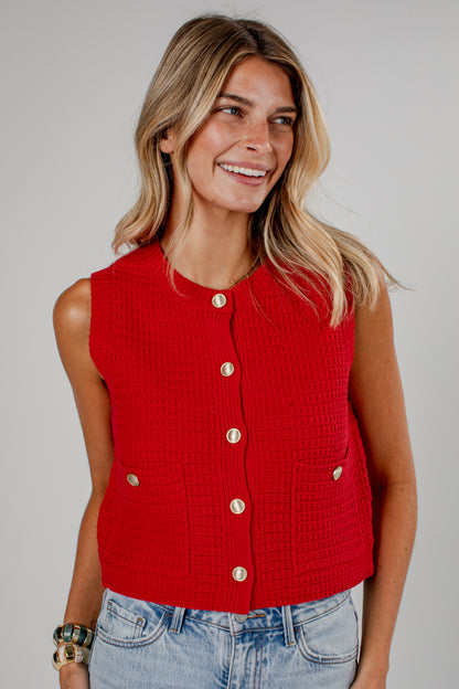 A woman wearing a bright red sleeveless cropped knit vest with gold-tone buttons and front pockets, paired with light-wash blue jeans, against a solid light gray studio background.