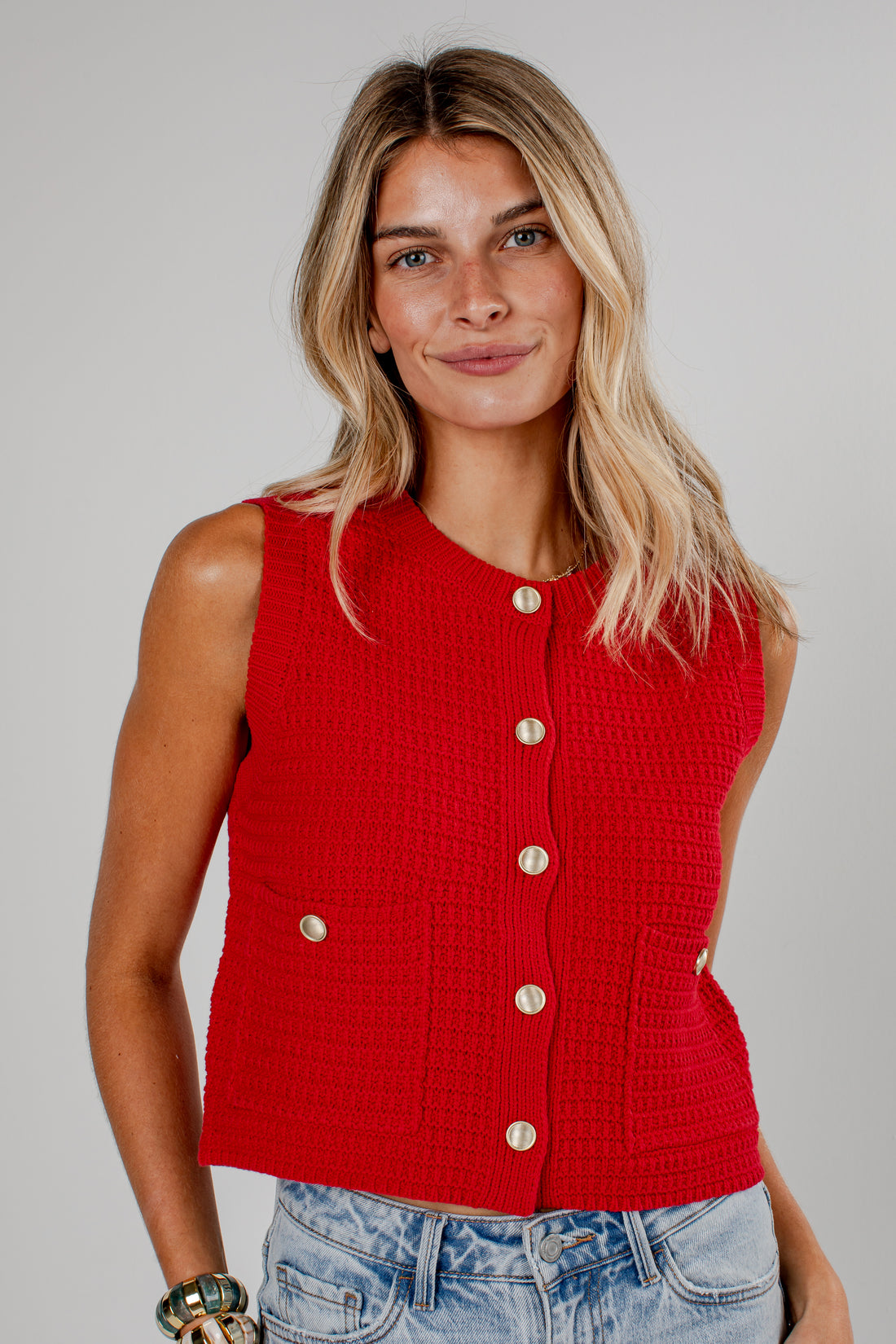A woman wearing a bright red sleeveless textured knit vest with gold-tone buttons, paired with light-wash blue jeans, against a solid light gray studio background.