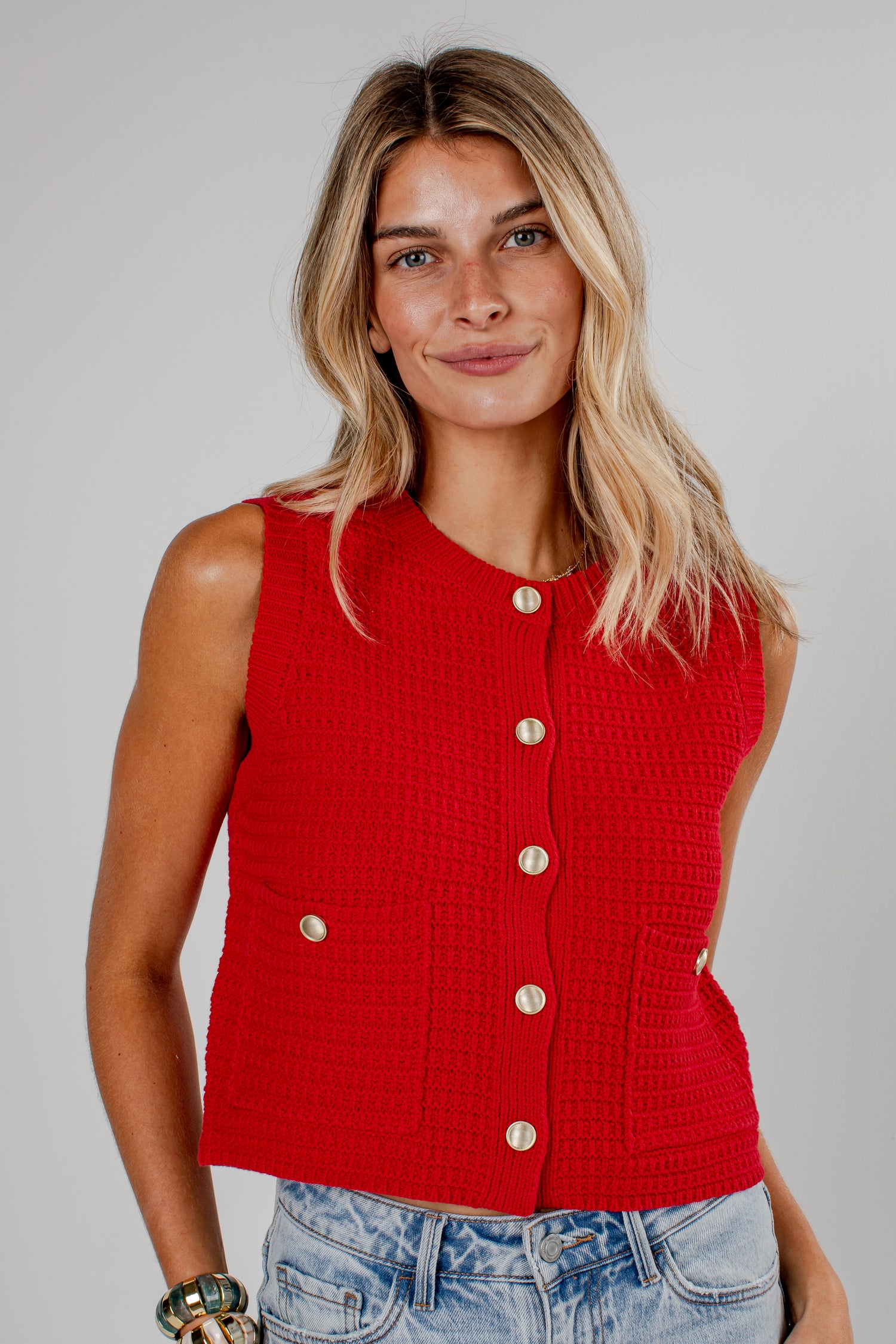 A woman wearing a bright red sleeveless textured knit vest with gold-tone buttons, paired with light-wash blue jeans, against a solid light gray studio background.