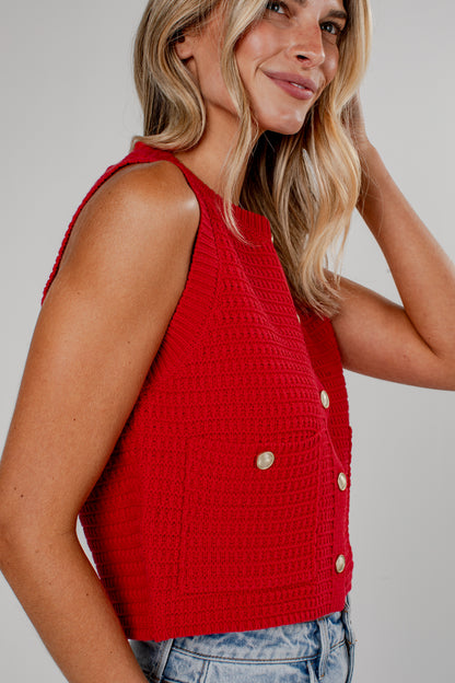 A woman wearing a bright red sleeveless cropped vest with a textured waffle knit pattern and decorative gold-tone buttons, paired with light-wash blue jeans against a neutral gray background.