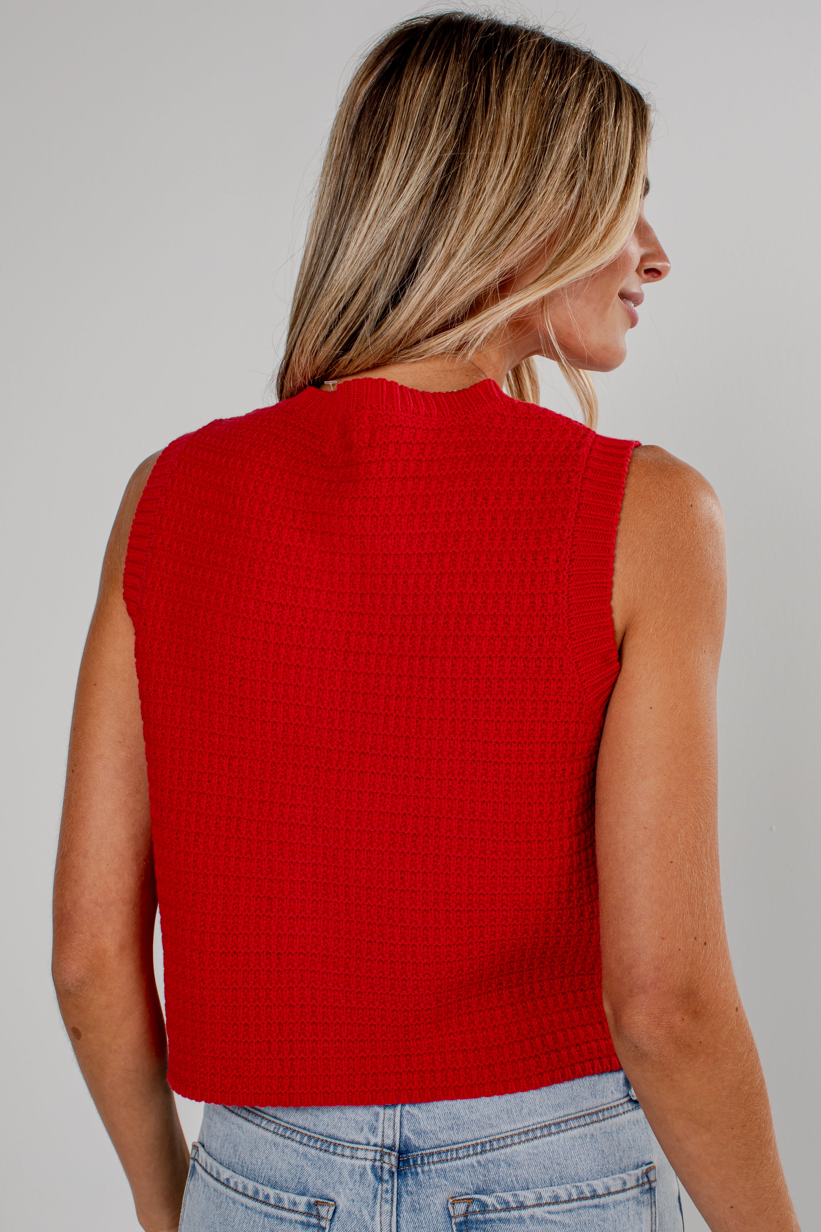 A woman shown from the back wearing a bright red sleeveless textured knit top and light blue jeans against a plain light gray studio background.