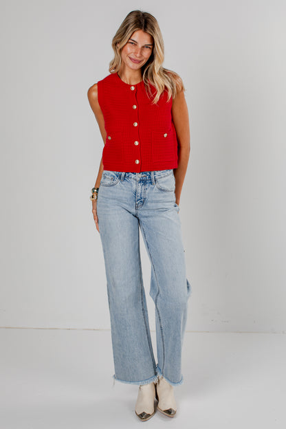 A woman modeling a sleeveless red knit vest with gold-tone buttons, paired with light-wash wide-leg jeans and beige pointed-toe heels, against a plain light gray studio background.