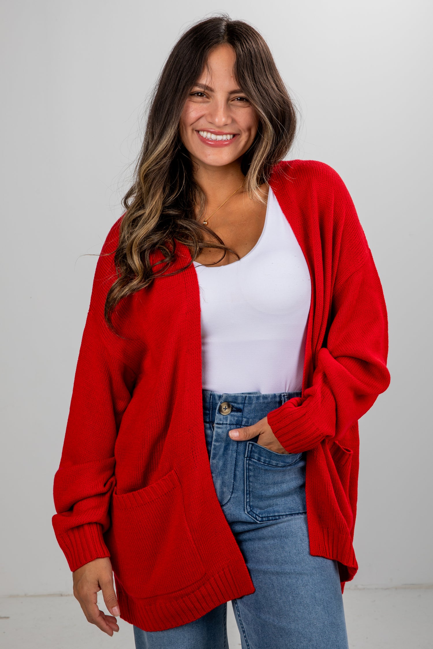 Woman wearing a red cardigan over a white top and blue jeans on a plain background