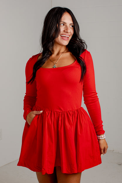 A woman modeling a bright red long-sleeve skater dress with a scoop neckline and side pockets, standing against a light gray wall.