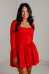 A woman modeling a bright red long-sleeve mini dress with a scoop neckline and a flared skirt, standing against a plain white wall.