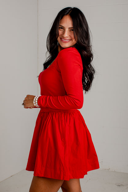 A woman with long dark hair wearing a bright red long-sleeve fit-and-flare dress, posing in a studio setting against a light gray wall.