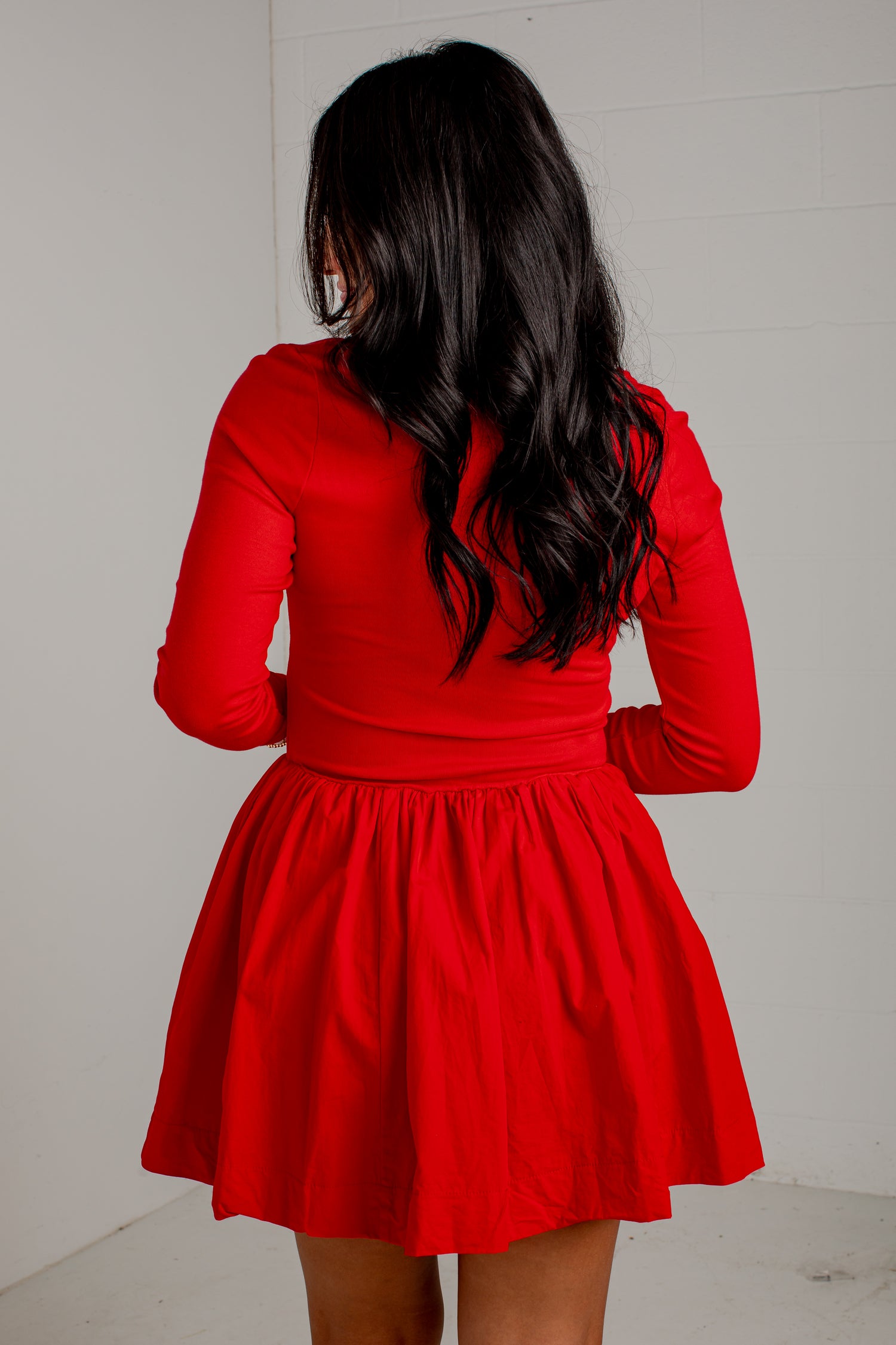 A woman viewed from behind wearing a bright red long-sleeve top and matching red flared skirt against a plain light gray wall.