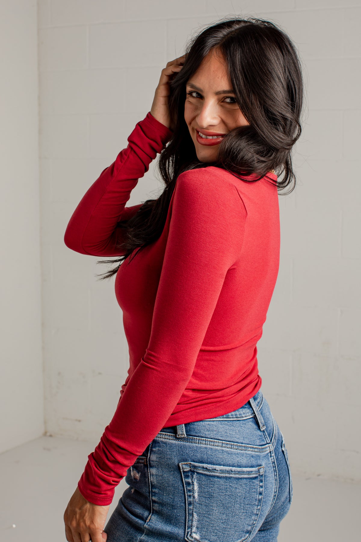 A woman with long dark hair wearing a fitted red long-sleeve top and blue jeans, posing in a studio setting against a white brick wall.