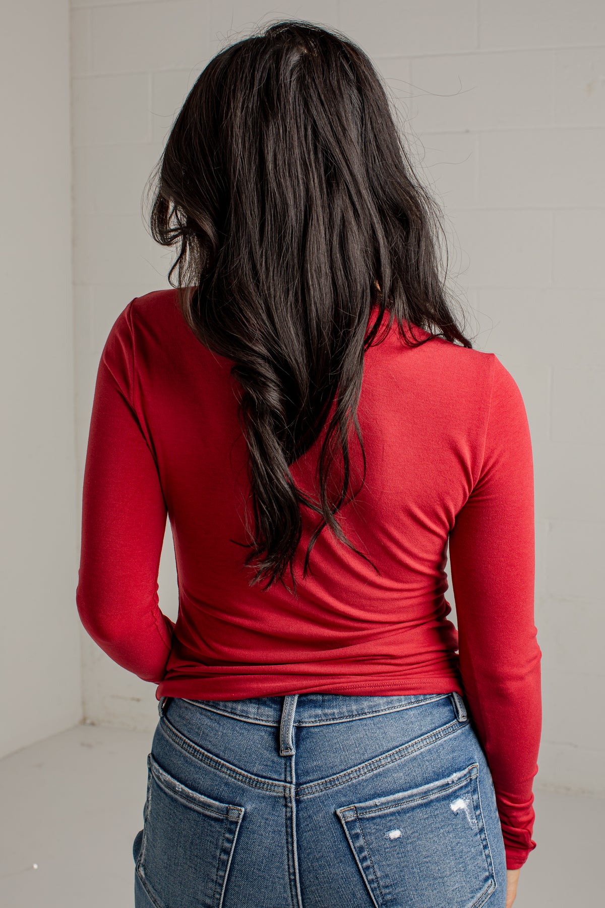 A woman viewed from behind wearing a fitted red long-sleeve top and blue jeans, standing against a plain light wall.