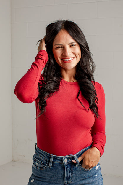 A smiling woman with long dark hair wearing a fitted red long-sleeve top and blue jeans, standing against a light gray wall.