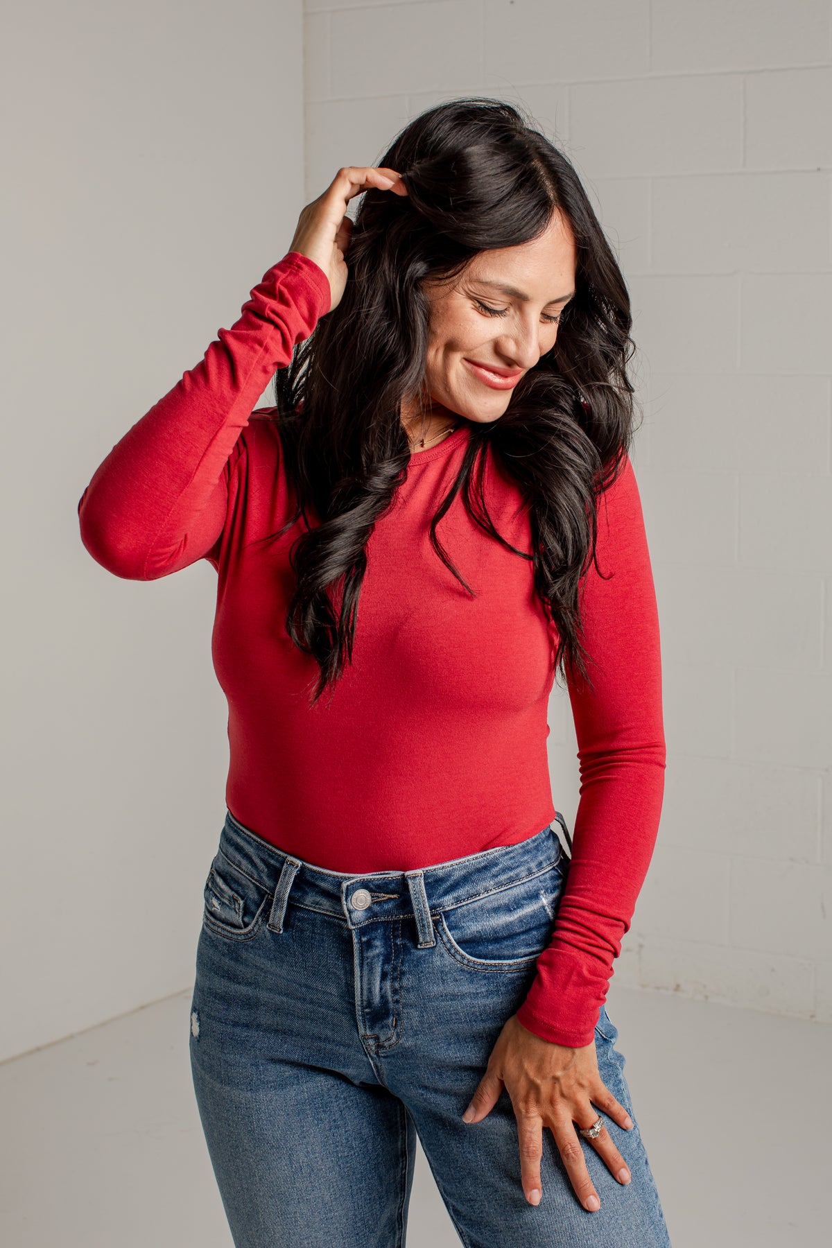 A woman with long dark hair wearing a fitted red long-sleeve top and blue jeans, standing against a light gray wall.