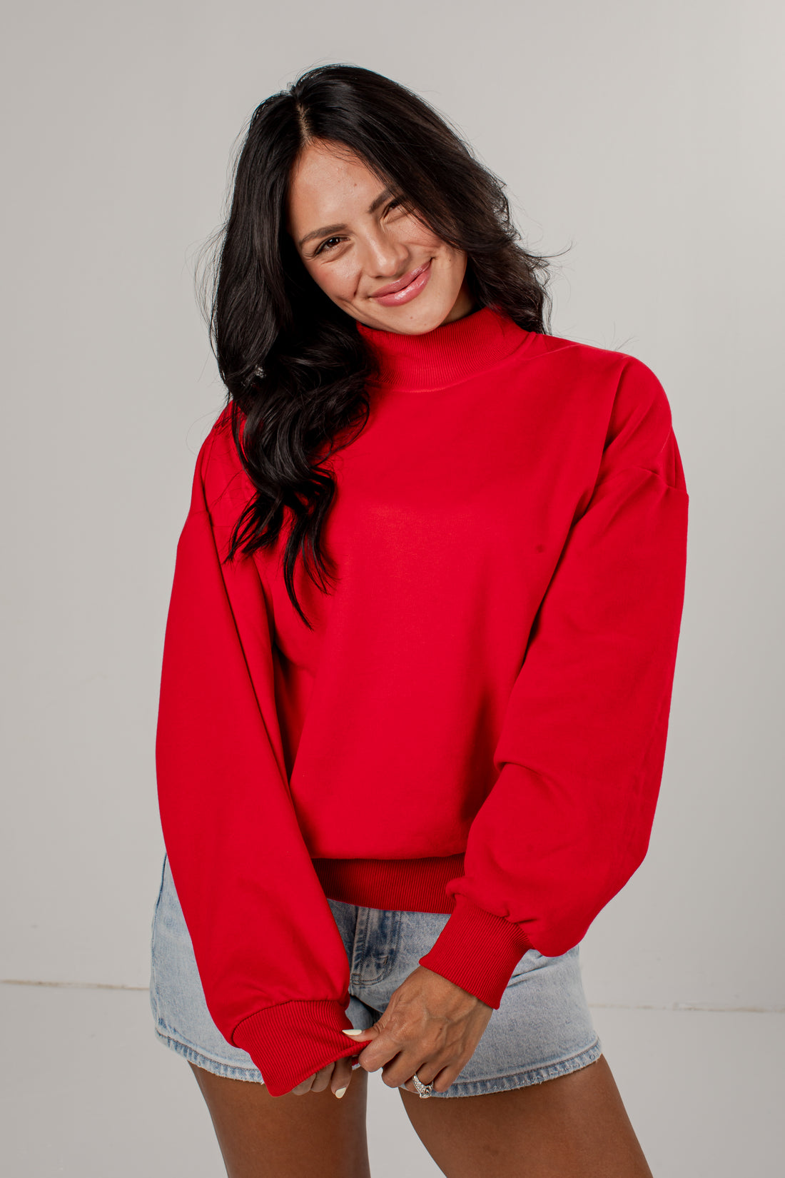 A woman wearing a bright red mock-neck sweatshirt and light-wash denim shorts, posing against a neutral light gray studio background.