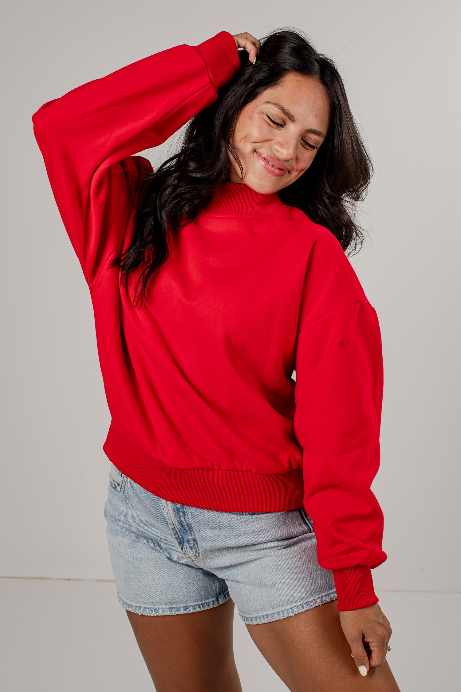 A woman wearing a bright red crewneck sweatshirt and light-wash denim shorts, posing against a neutral light gray studio background.