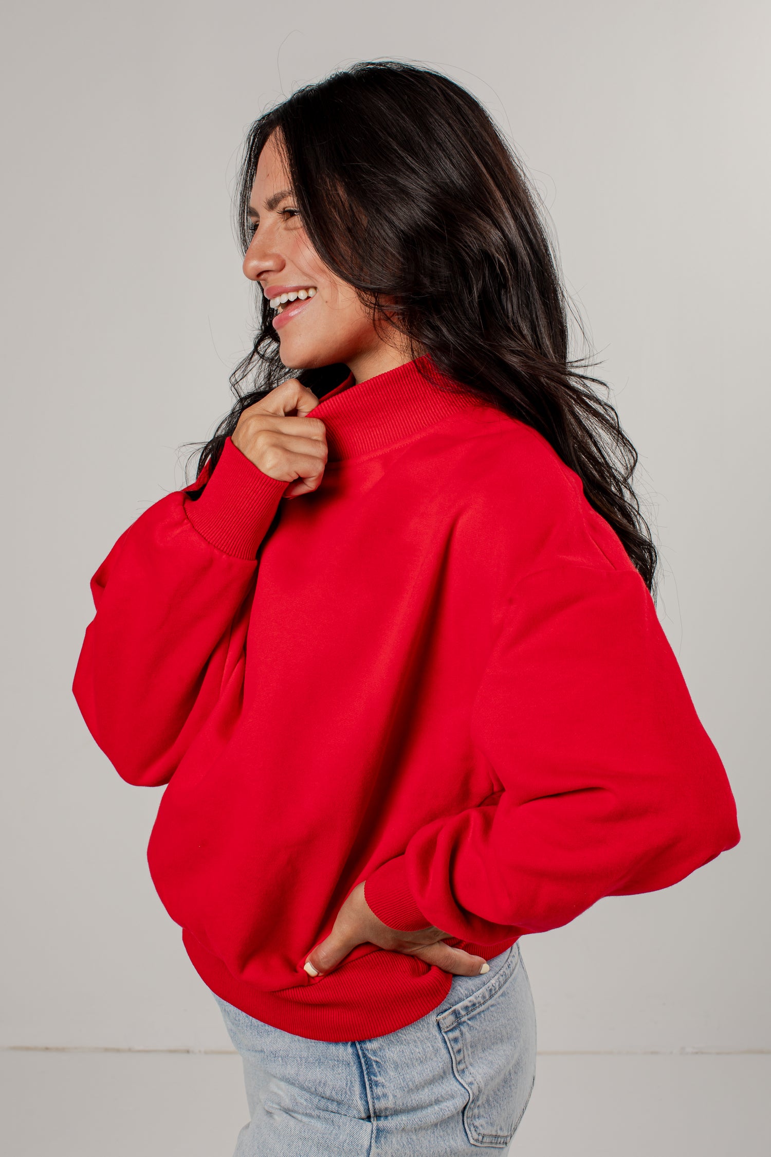 A woman wearing a bright red crewneck sweatshirt and light blue jeans, shown from a side profile against a plain light gray studio background.