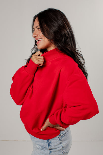 A woman wearing a bright red crewneck sweatshirt and light blue jeans, shown from a side profile against a plain light gray studio background.