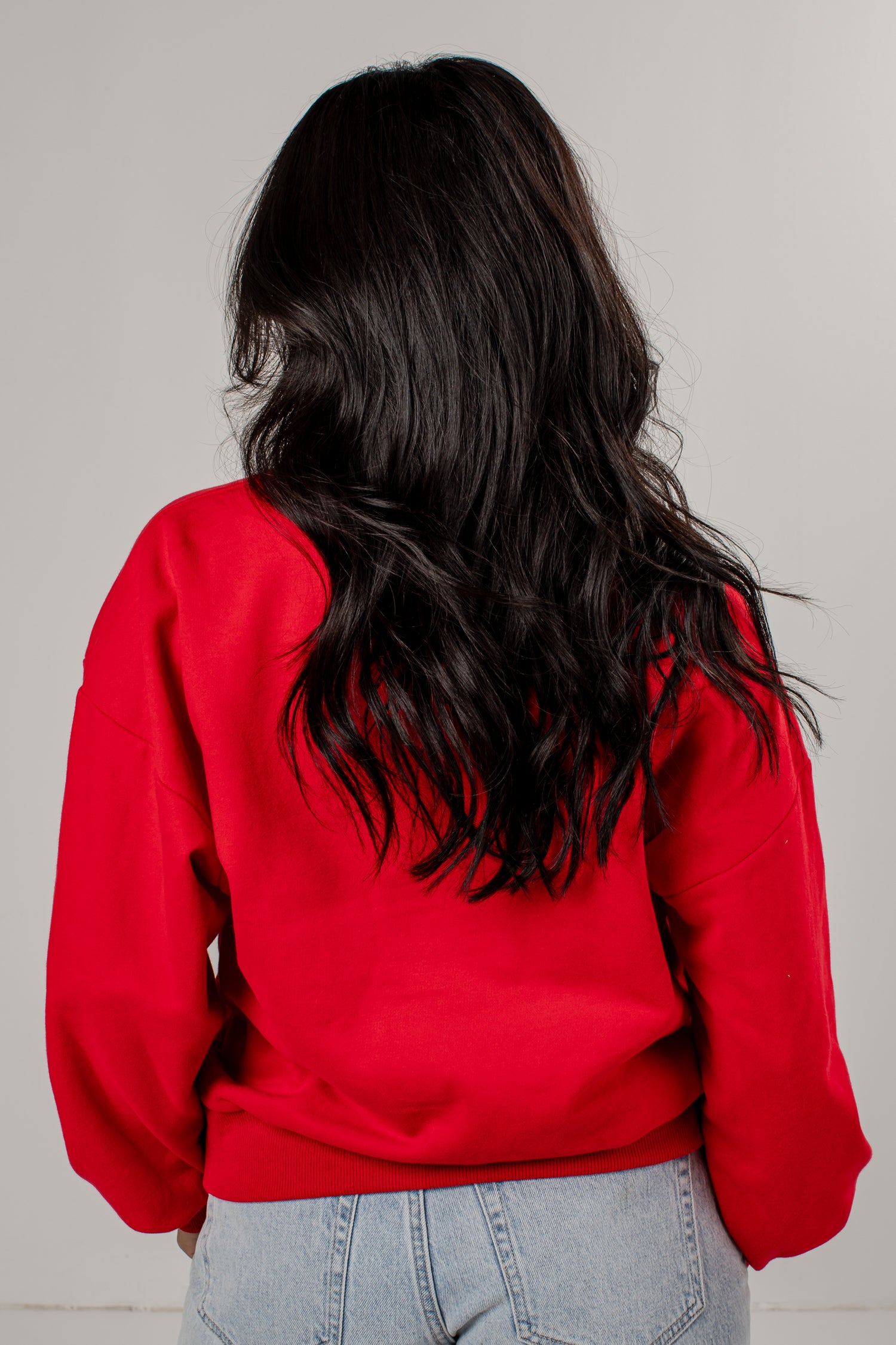 A person viewed from behind wearing a bright red crewneck sweatshirt and light blue jeans, with long dark wavy hair, against a plain light gray background.