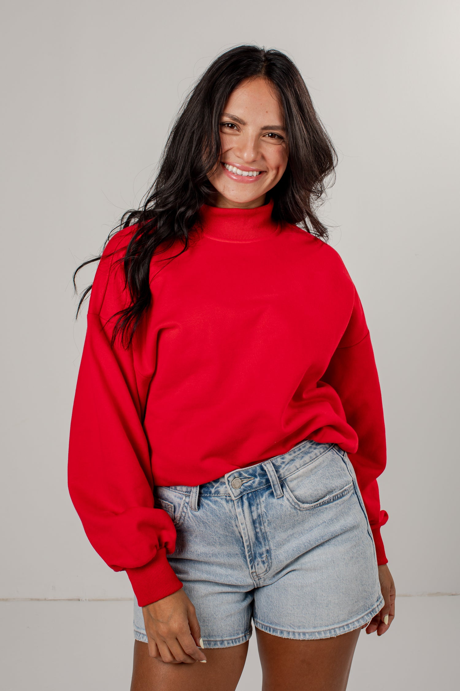 A woman with long dark hair wearing a bright red mock-neck sweatshirt and light-wash denim shorts, standing against a neutral light gray studio background.