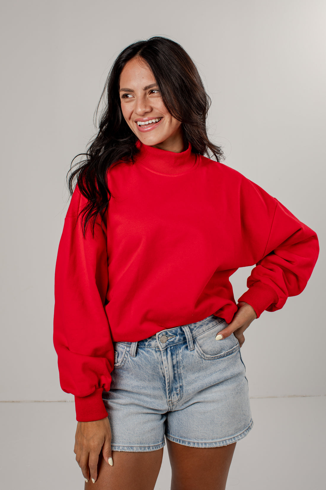 A woman wearing a bright red mock-neck sweatshirt and light-wash denim shorts, standing against a plain light gray studio background.