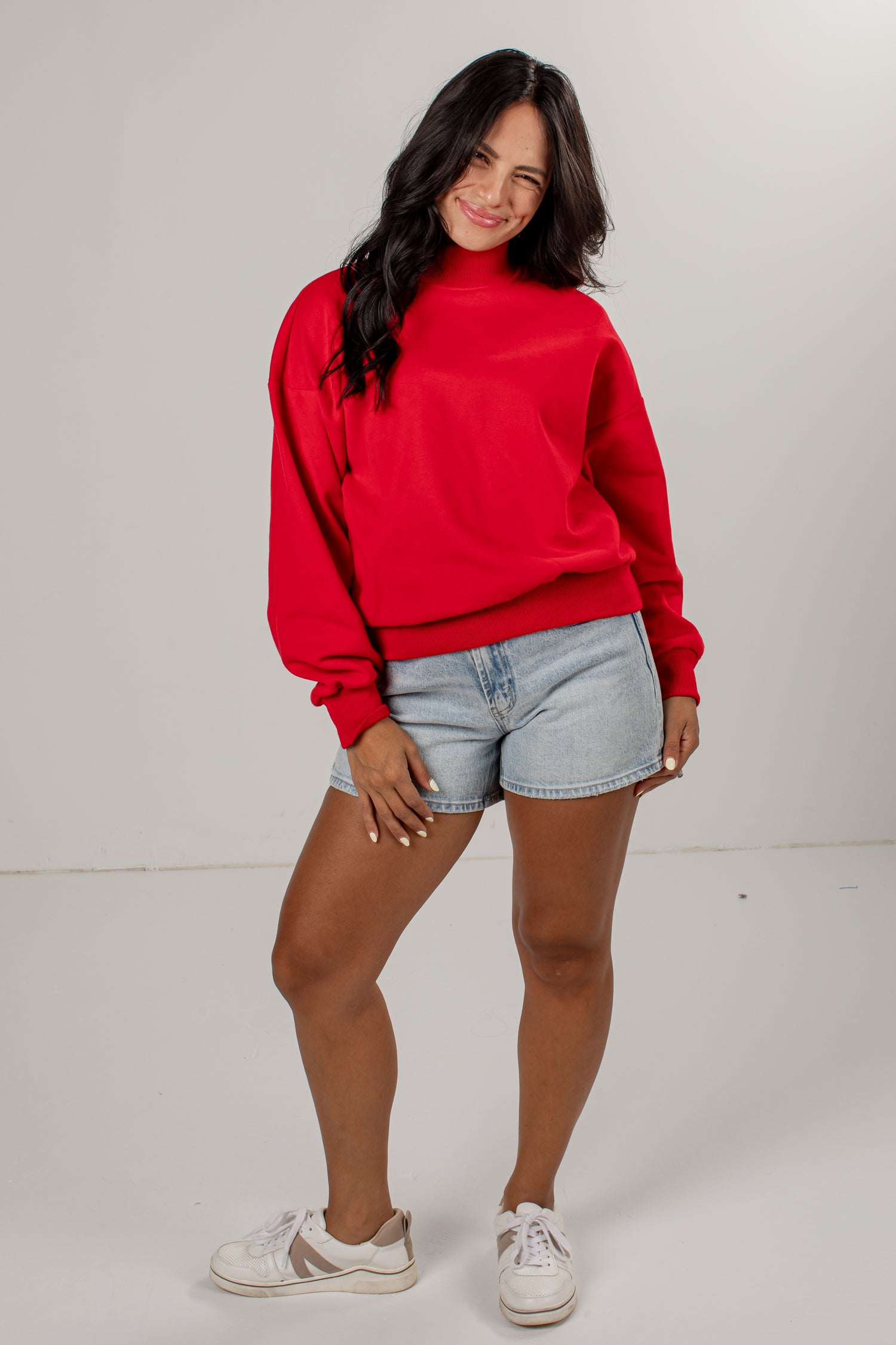 A woman modeling a bright red long-sleeve sweatshirt, paired with light-wash denim shorts and white sneakers, standing against a plain light gray studio background.