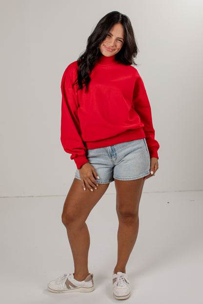 A woman modeling a bright red long-sleeve sweatshirt, paired with light-wash denim shorts and white sneakers, standing against a plain light gray studio background.