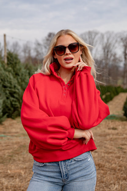 Woman wearing a red hoodie and sunglasses in an outdoor setting with trees and grass.