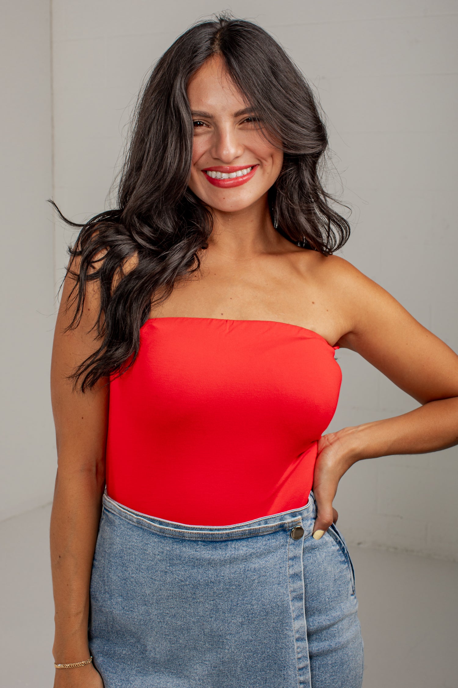 A woman with long dark hair wearing a bright red strapless tube top and a light blue denim skirt, posing against a neutral light gray background.