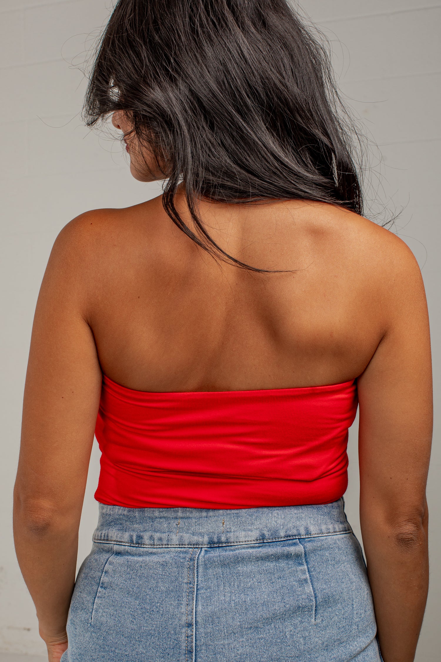 A woman shown from behind wearing a bright red strapless bandeau top and light blue denim jeans against a plain light gray wall.