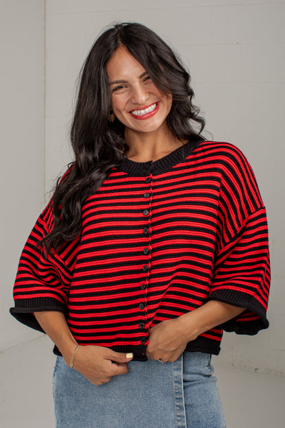 A smiling woman with long dark hair wearing a red and black horizontally striped knit top with wide dolman sleeves and a button front, paired with light blue jeans, standing against a light gray wall.