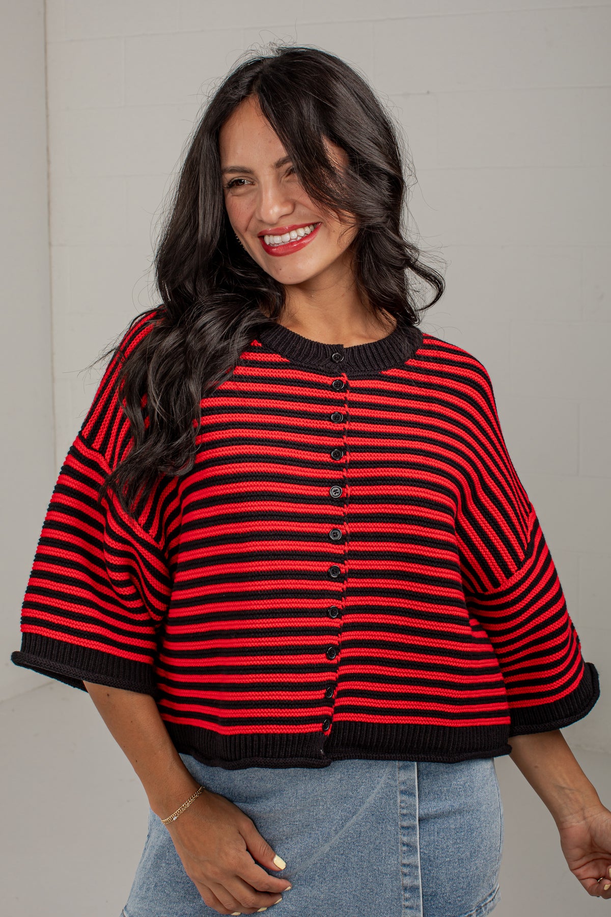 A woman wearing a red and black striped knit cardigan with wide three-quarter sleeves and a button-front closure, paired with light blue jeans against a light gray wall.