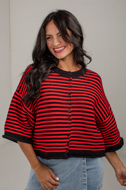 A woman wearing a red and black striped knit cardigan with wide three-quarter sleeves and a button-front closure, paired with light blue jeans against a light gray wall.